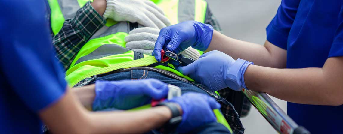 Persoa colocada nunha padiola tras sufrir un accidente laboral. A persoa está situado no centro da imaxe, acostado e leva ropa de traballo. Ao carón, a cada lado hai unha persoa do sector da saúde prestando coidados
