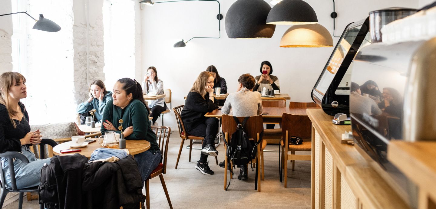 Interior dunha cafetería, na dereita da imaxe pode verse o mostrador e na parte dereita da imaxe, hai varias mesas con xente sentada a falar.