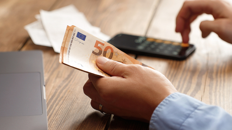 Na imaxe vense a mans dunha persoa, na man esquerda leva varios billetes de 50 euros e coa man dereita está a utilizar unha calculadora
