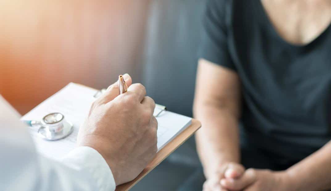 Persoa sendo atendida nunca consulta médica. No primeiro plano está o persoal sanitario tomando notas e fronte a el a persoa que está a ser atendida.